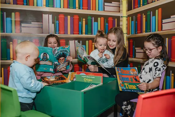 An adult reads picture books to four young children seated around a green table in a library with colorful books on shelves behind them.