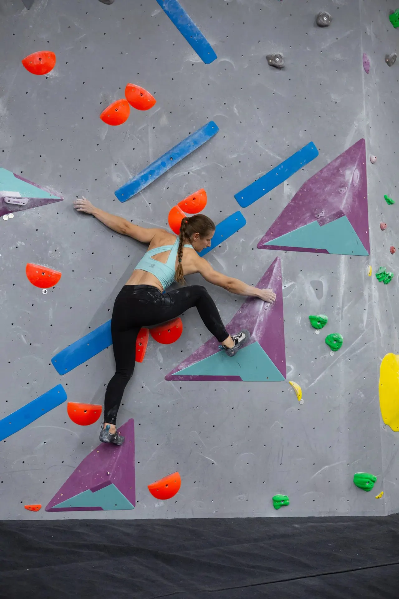 Woman climbing an indoor rock wall with coloured handholds and geometric shapes.
