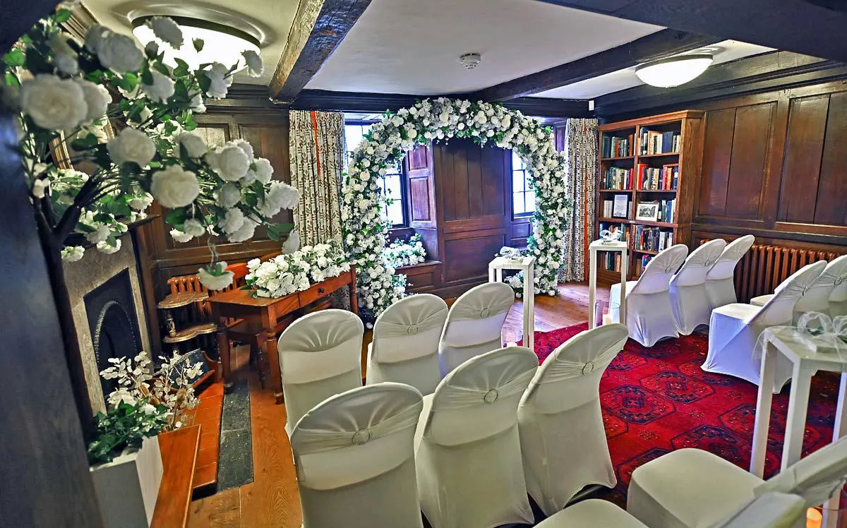 Elegant interior ceremony room with white-covered chairs facing a floral arch, surrounded by white flowers and lush green garlands.