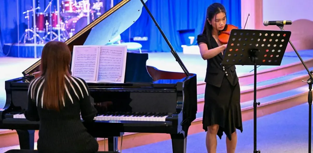 Two young musicians perform on stage under blue and pink lighting; one plays grand piano while the other stands with a violin, reading from sheet music on a stand.