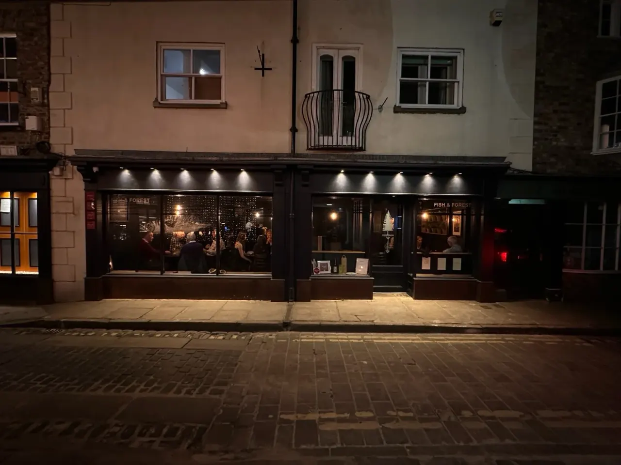 Illuminated restaurant storefront with black shopfronts and windows on a night-time street with diners visible inside.