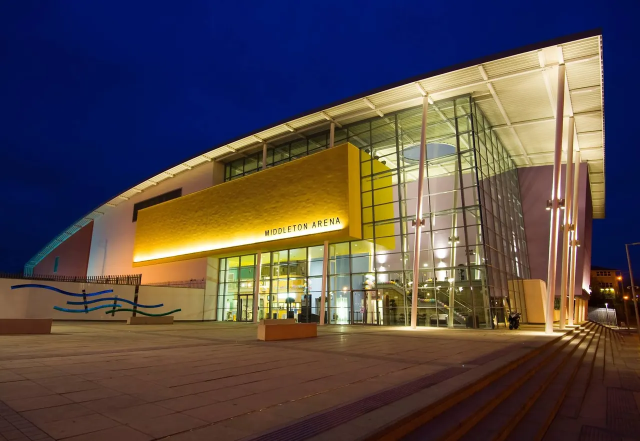 Modern illuminated arena building with yellow signage at dusk against a deep blue sky.