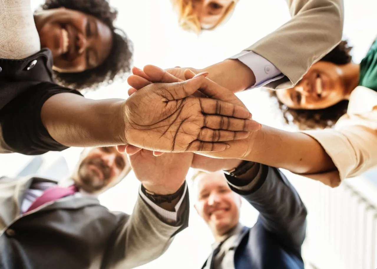 A group of children and adults place their hands together in a unified stack, viewed from below against a bright sky.