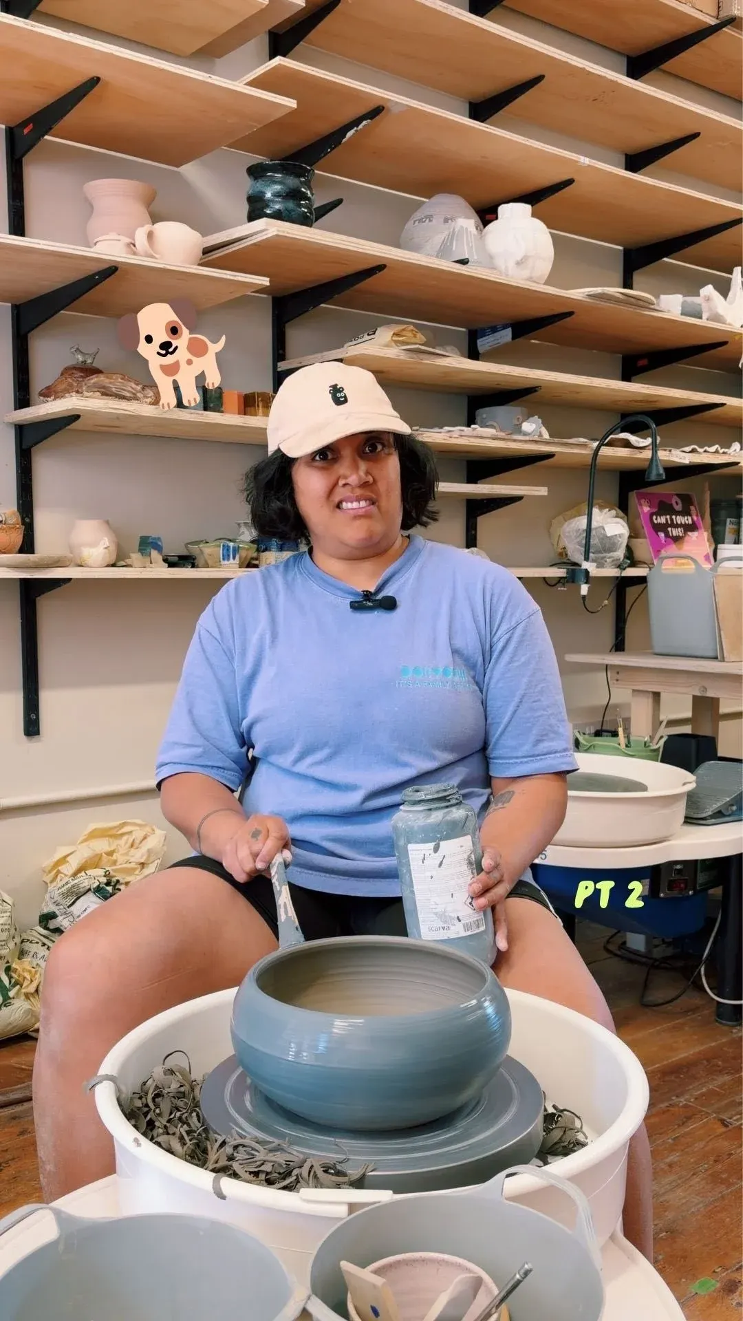 Person at pottery wheel demonstrating ceramic making in a studio with shelved displays of finished pottery pieces.