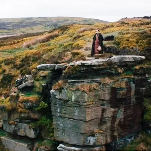 Person standing on rocky outcrop in moorland with rolling hills in background.