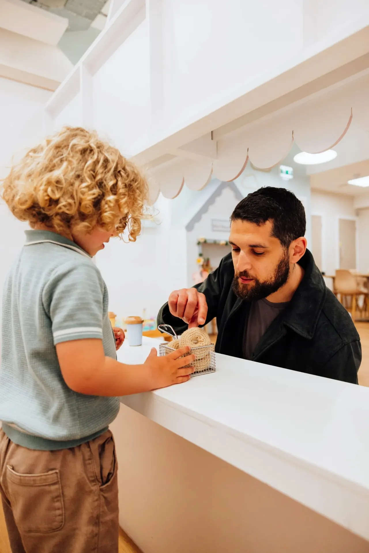 A young child with curly hair hands a small shopping basket to an adult man across a white counter in a bright, modern play café interior.