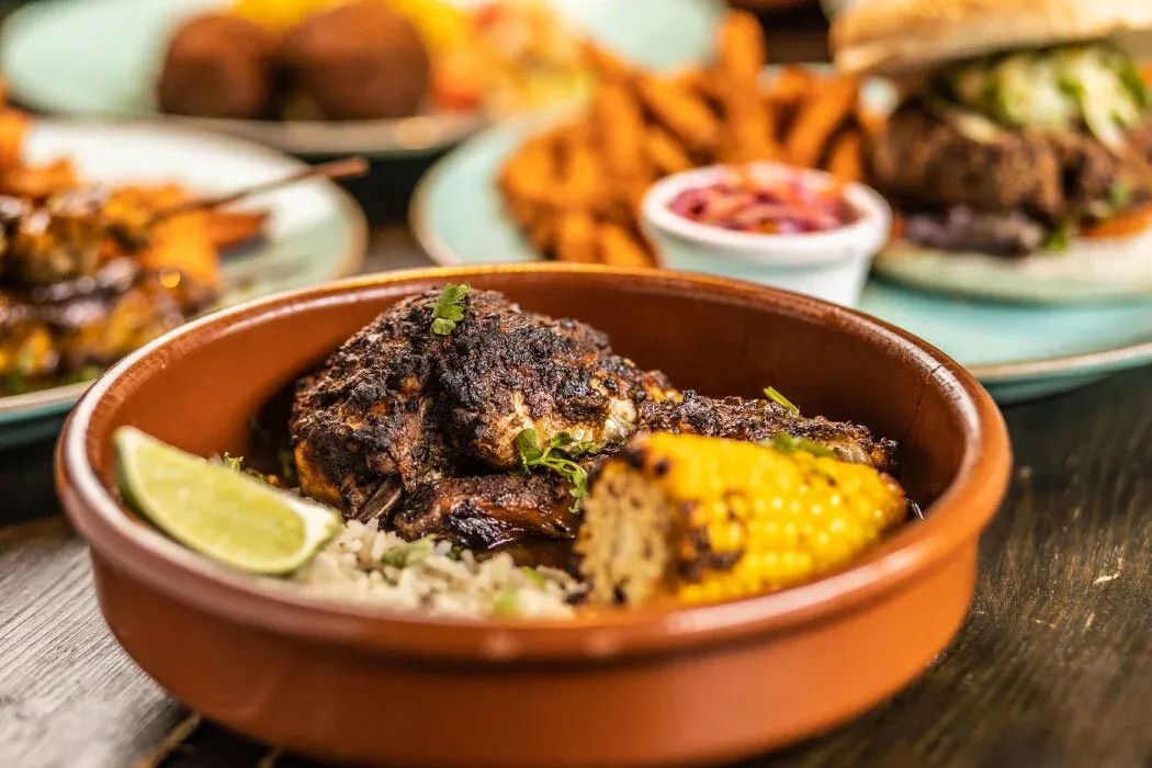 Grilled meat with corn, lime and rice in a clay bowl, with other dishes blurred behind.