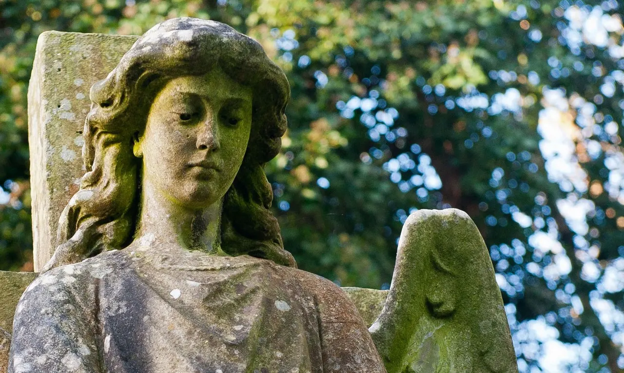 Stone statue of a woman with long hair and contemplative expression, covered in moss, surrounded by green foliage.