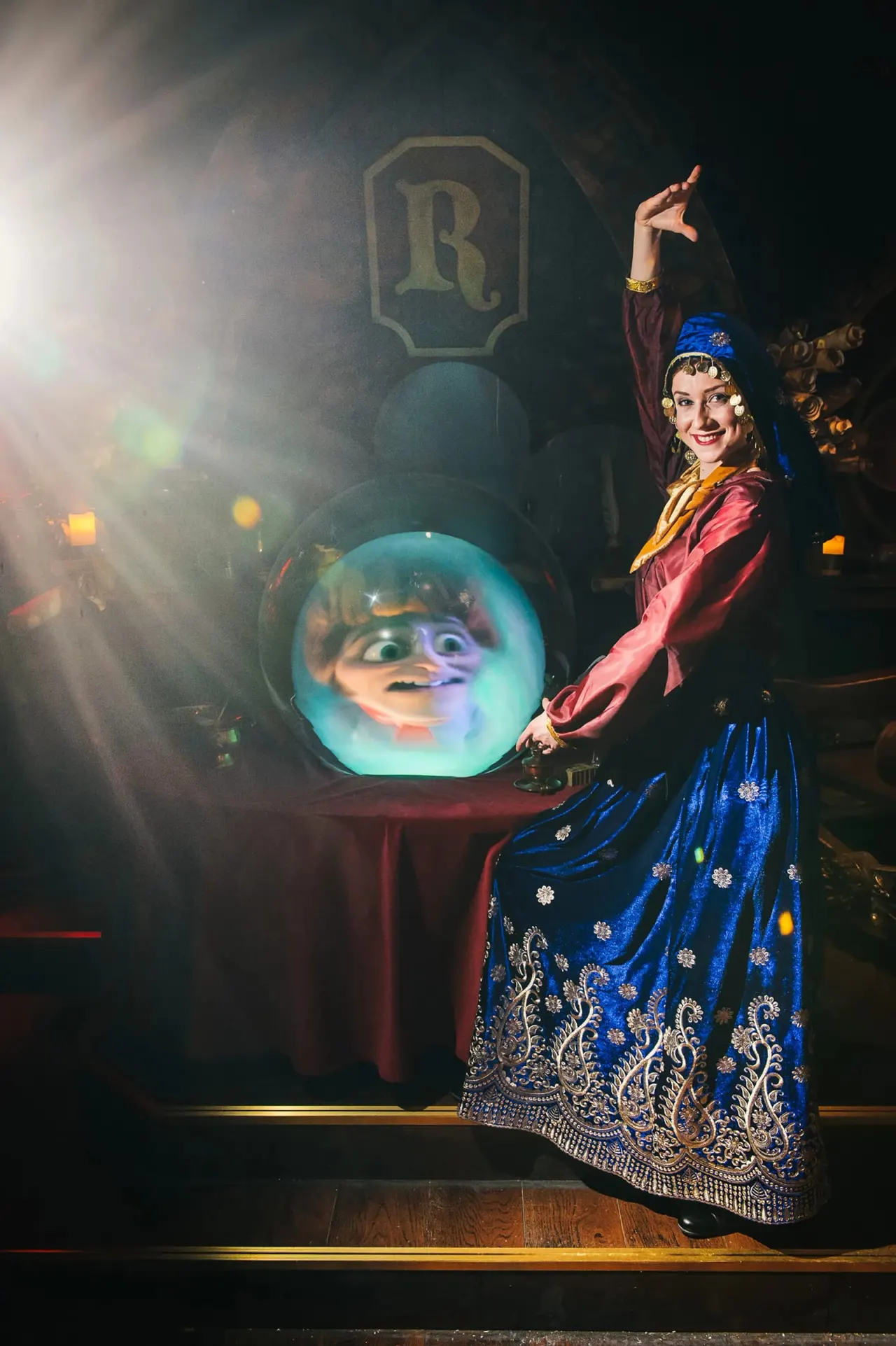 Performer in theatrical costume poses beside an illuminated crystal ball displaying an animated character's face during an indoor attraction show.