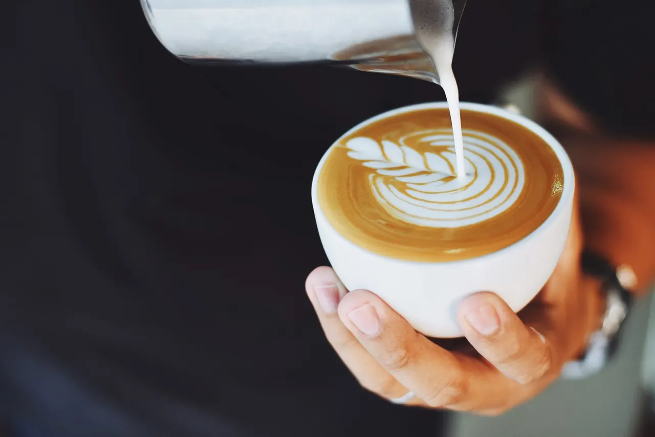 Hand holding white cup of latte with latte art while steamed milk is being poured in.