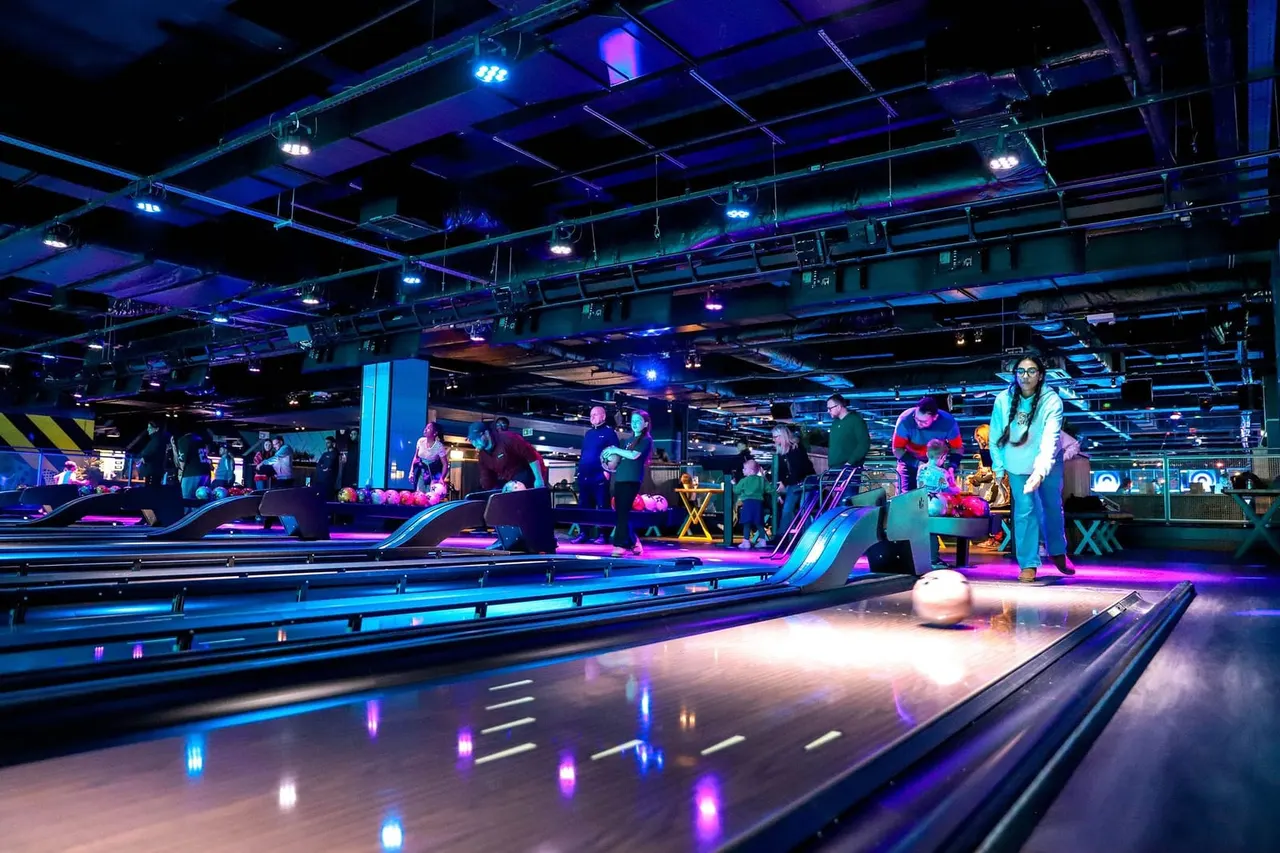 Indoor tenpin bowling alley with neon lighting, multiple lanes, and visitors of various ages bowling and socialising.
