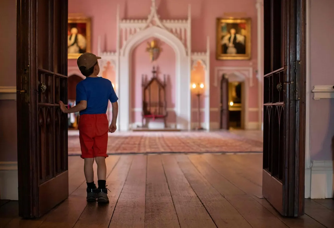 Child in blue shirt and red shorts standing in grand pink palace corridor with ornate doorways and portraits.
