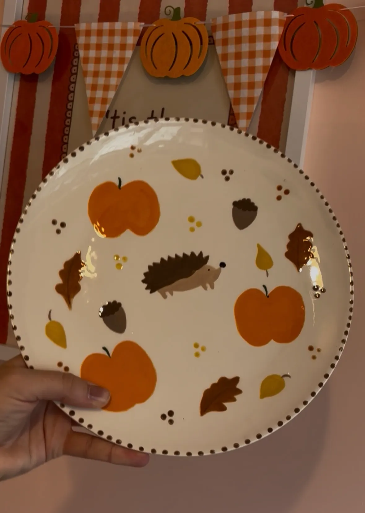 Hand holding a decorative autumn-themed plate with apples, leaves and hedgehog illustrations, displayed against striped backdrop with hanging pumpkin decorations.