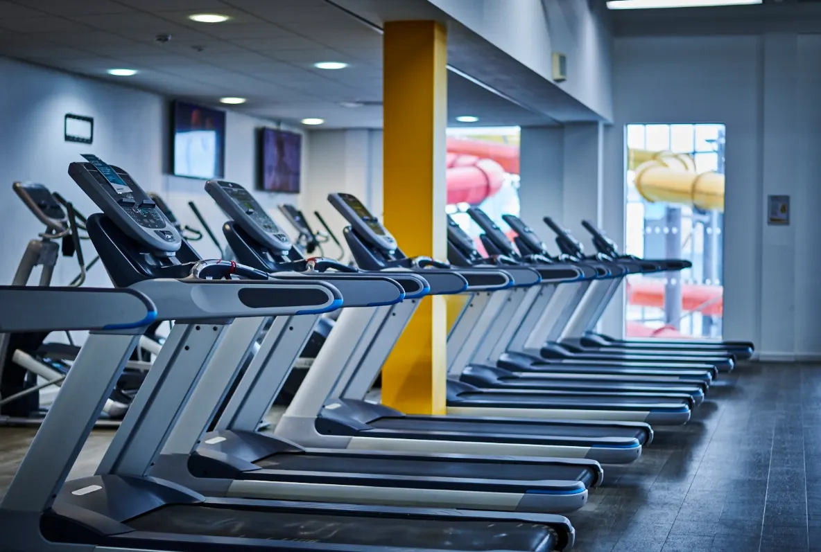 Row of treadmills in a modern gym with yellow support pillar and bright windows.