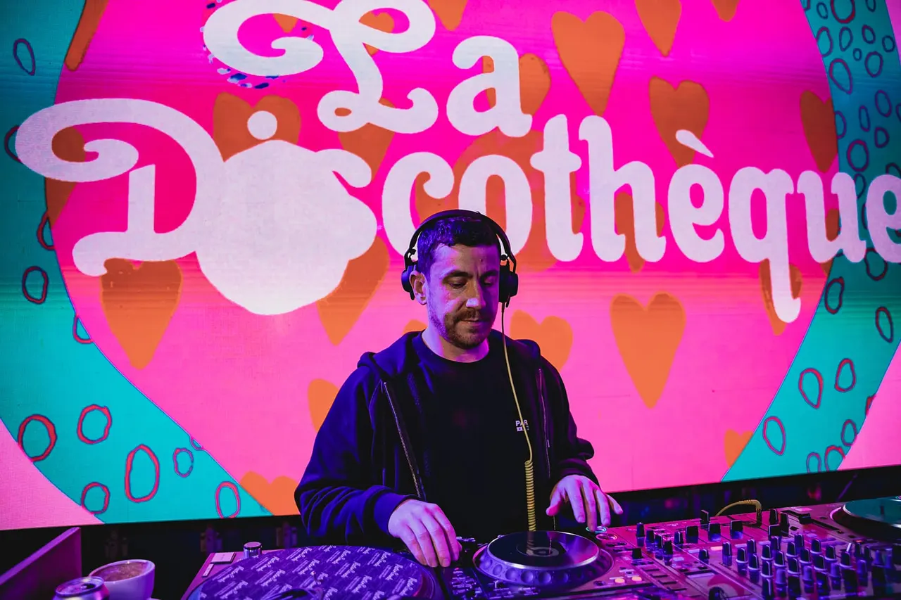 DJ wearing headphones at turntables performing in front of a vibrant pink and orange illuminated backdrop with hearts.