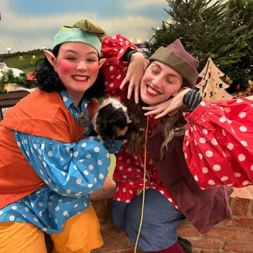 Two smiling staff members in colourful costumes hold a small dog while posing outdoors at a festive venue.