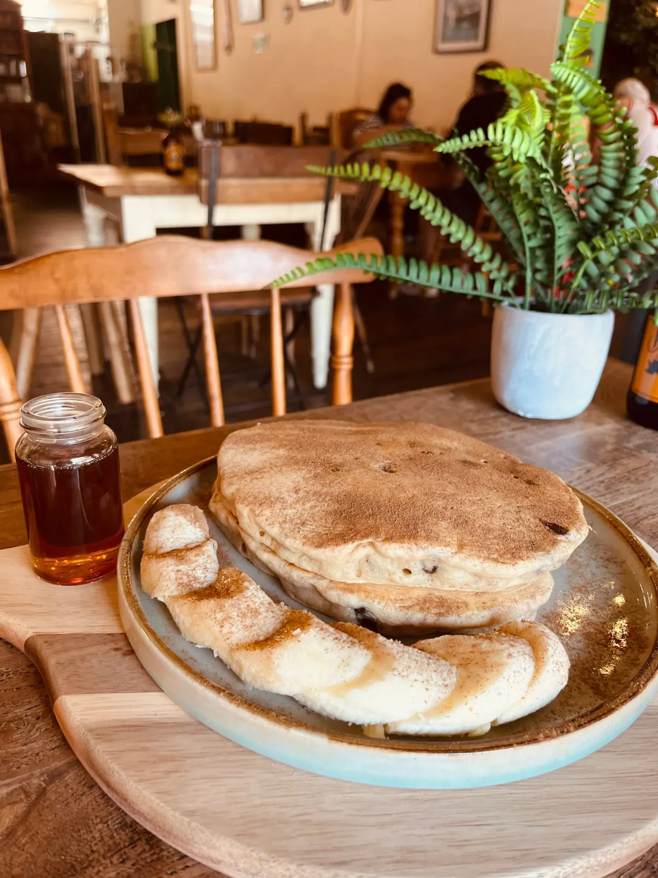 Plate of cinnamon-dusted pancakes and pastries with syrup on a café table, other diners visible in background.