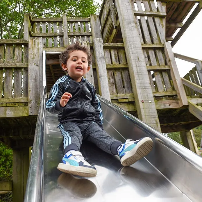 Young child sliding down a metal chute attached to a wooden play structure outdoors.