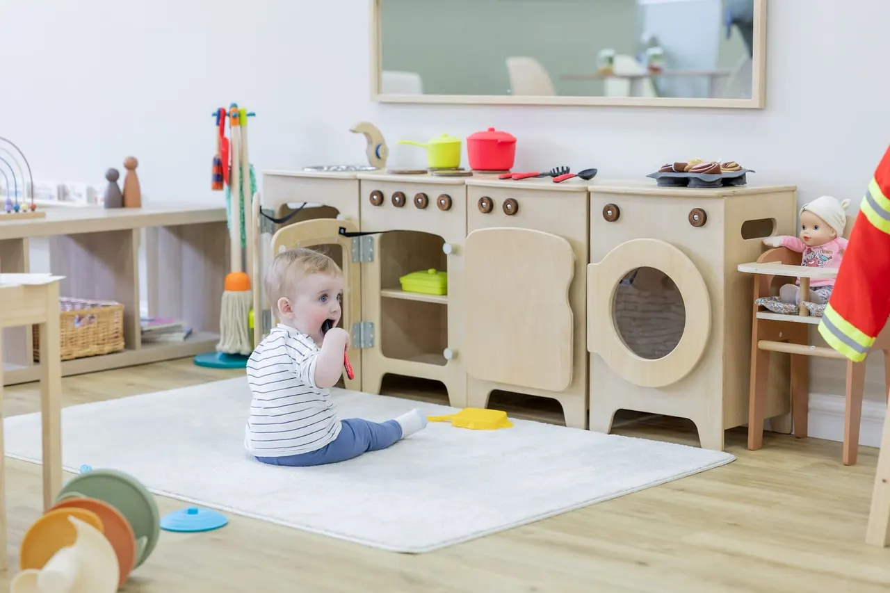A toddler in a striped shirt plays in a bright, wooden play kitchen area with toy appliances and cookware.