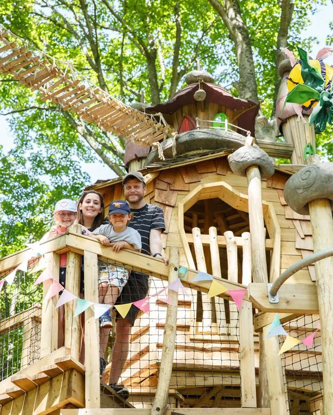 Family of four standing on wooden treehouse platforms amongst tall trees with colourful bunting decorations.