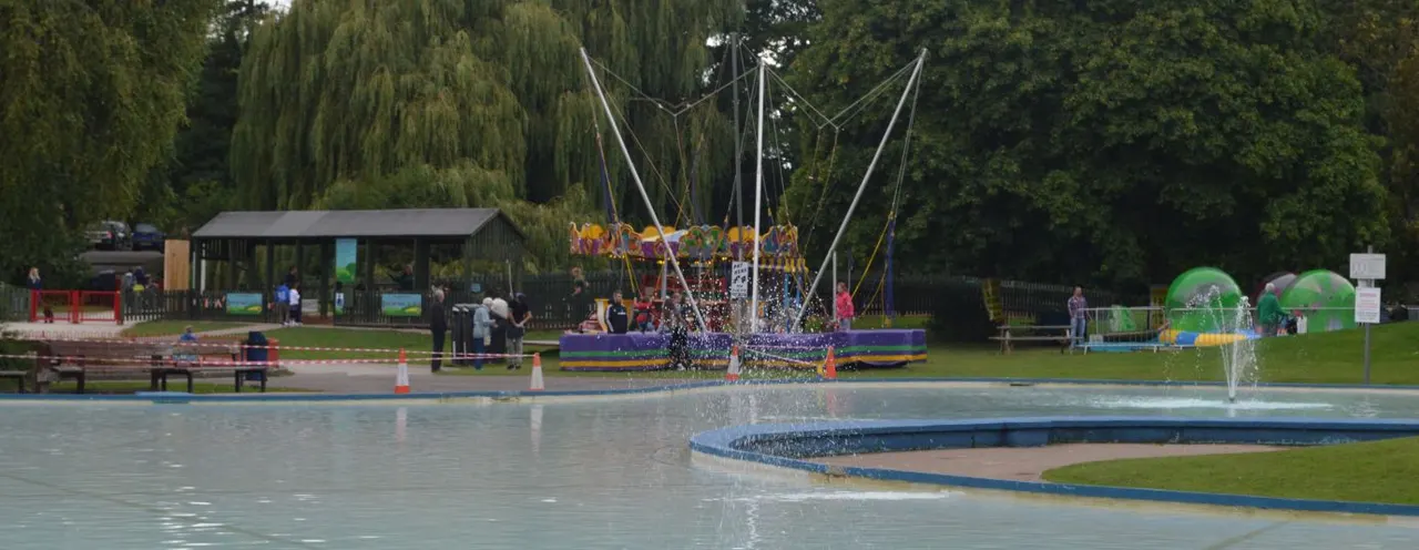 Outdoor water play area with shallow pools, a large colourful water-spraying play structure, and green inflatables, surrounded by trees and grassy grounds.