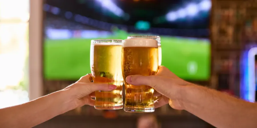 Two people toasting with pints of beer whilst watching a football match on screen.