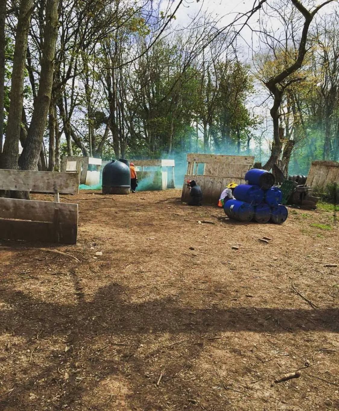 Paintball players in blue and orange gear take cover behind tyres and wooden barriers during a game in a wooded arena.