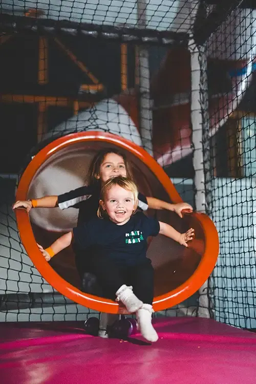 Two children playing inside a large orange tunnel on a pink bouncy surface within an indoor play area.