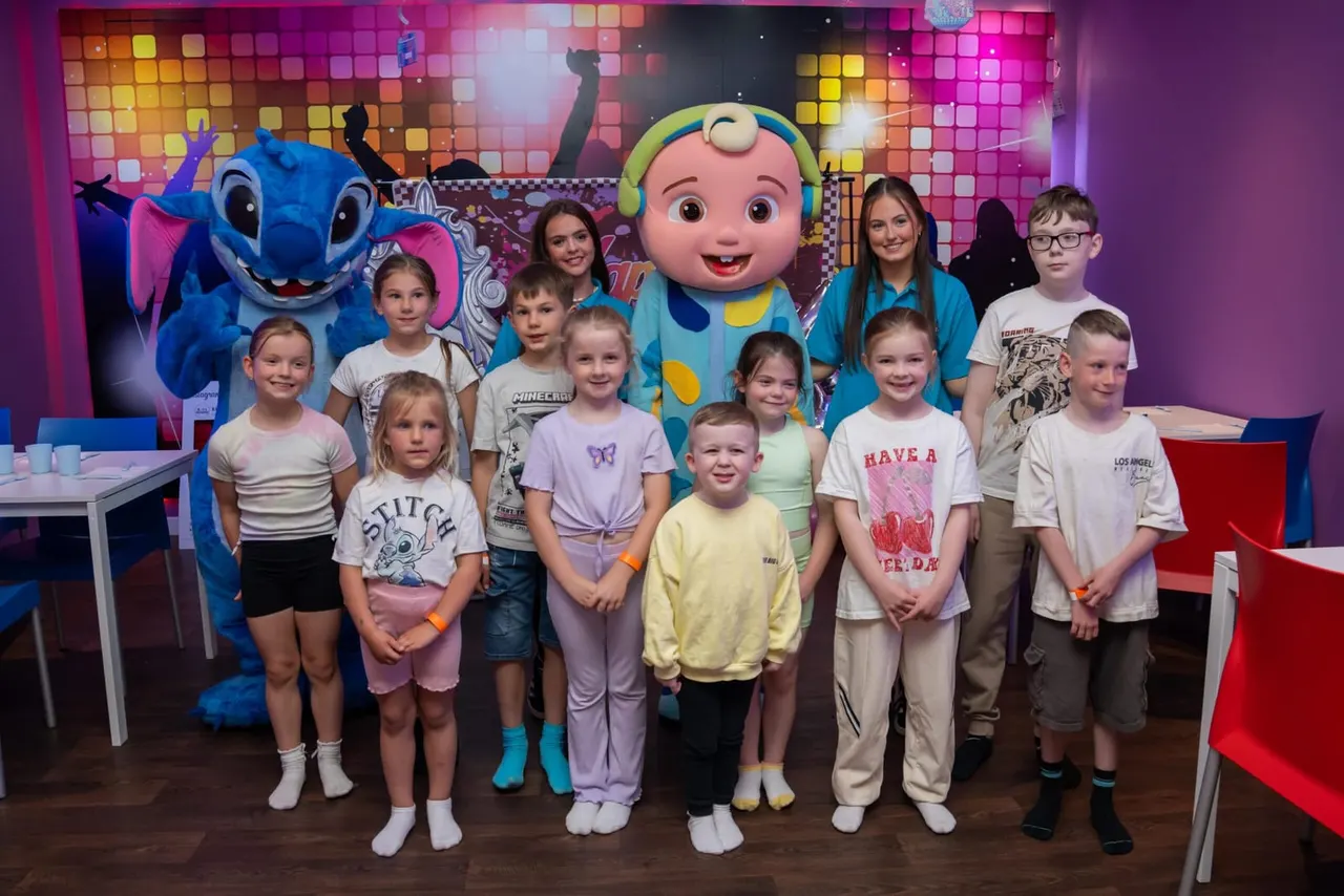 Group of children posing with costumed characters in a colourful indoor entertainment space with neon lighting.