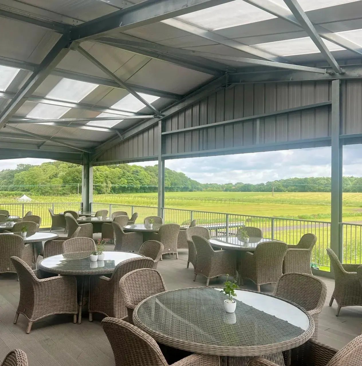 Covered outdoor seating area with wicker chairs and tables overlooking open green fields and woodland.