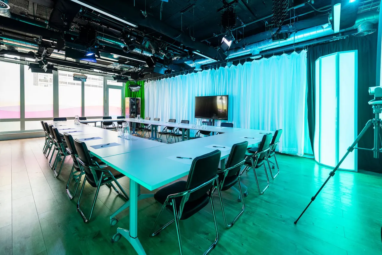 Modern conference room with turquoise lighting, long meeting tables, black chairs, screen display, and floor-to-ceiling windows.