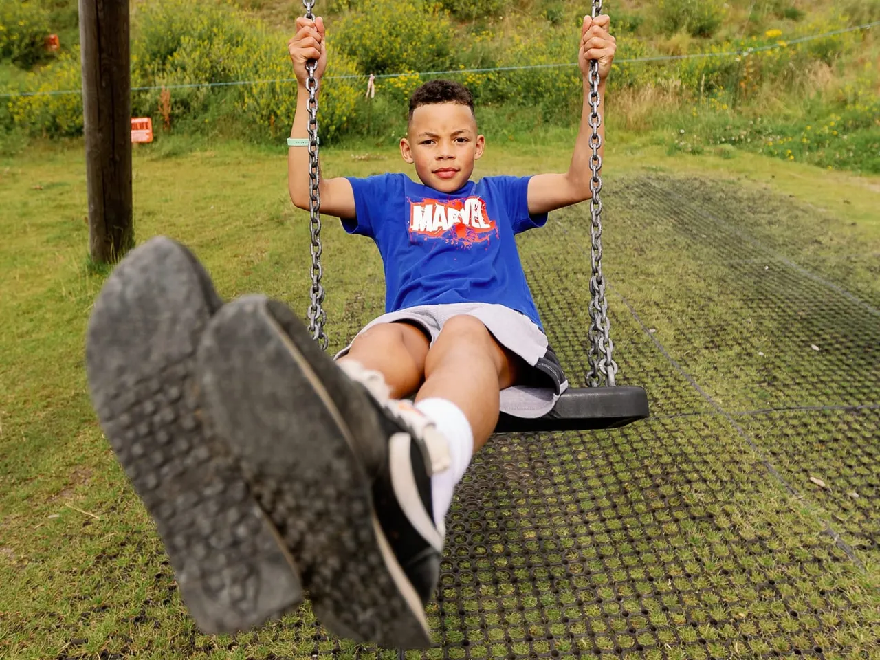 Child on a chain swing in a grassy outdoor play area with vegetation in background.