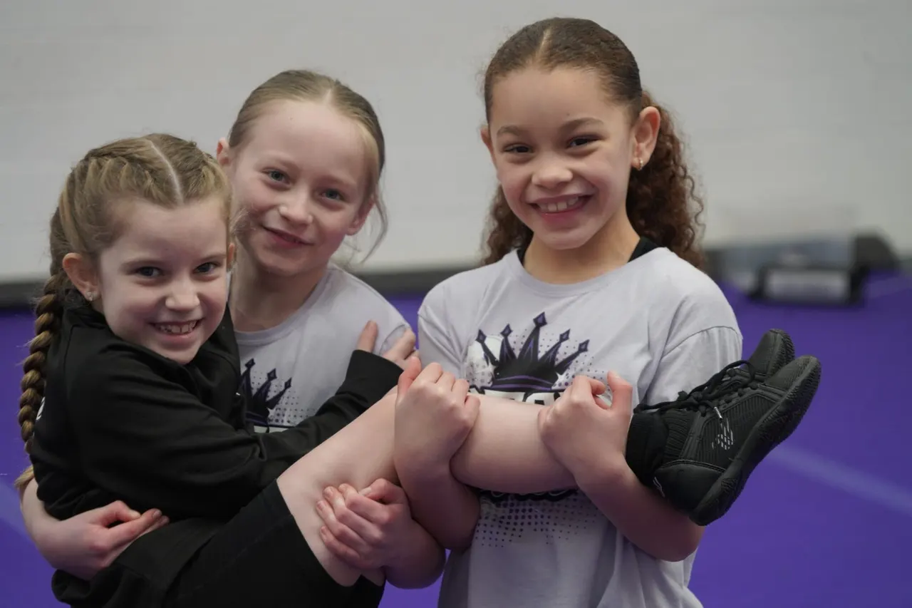 Three smiling girls in sports clothing posing together on a purple gymnastics mat.