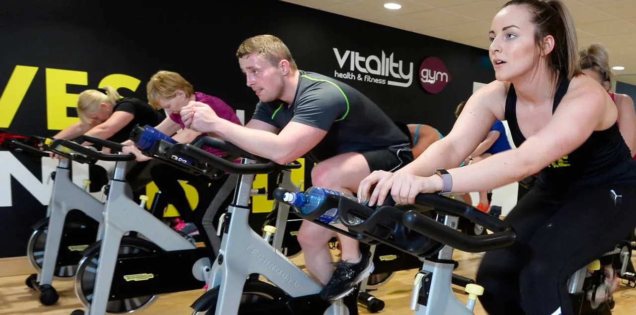Group of people on stationary bikes during a spin class at a gym with Vitality branding visible.