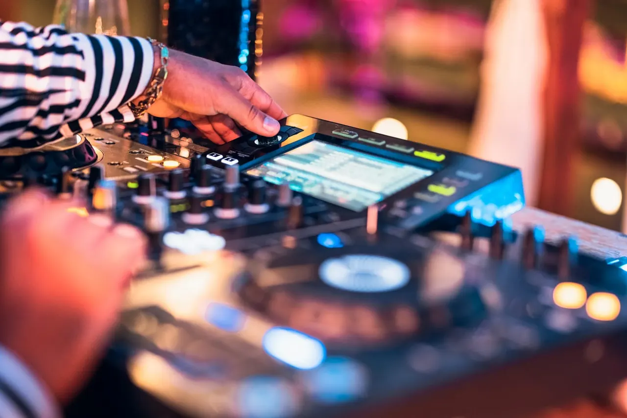 DJ's hand operating electronic mixing equipment with colourful stage lighting in the background.