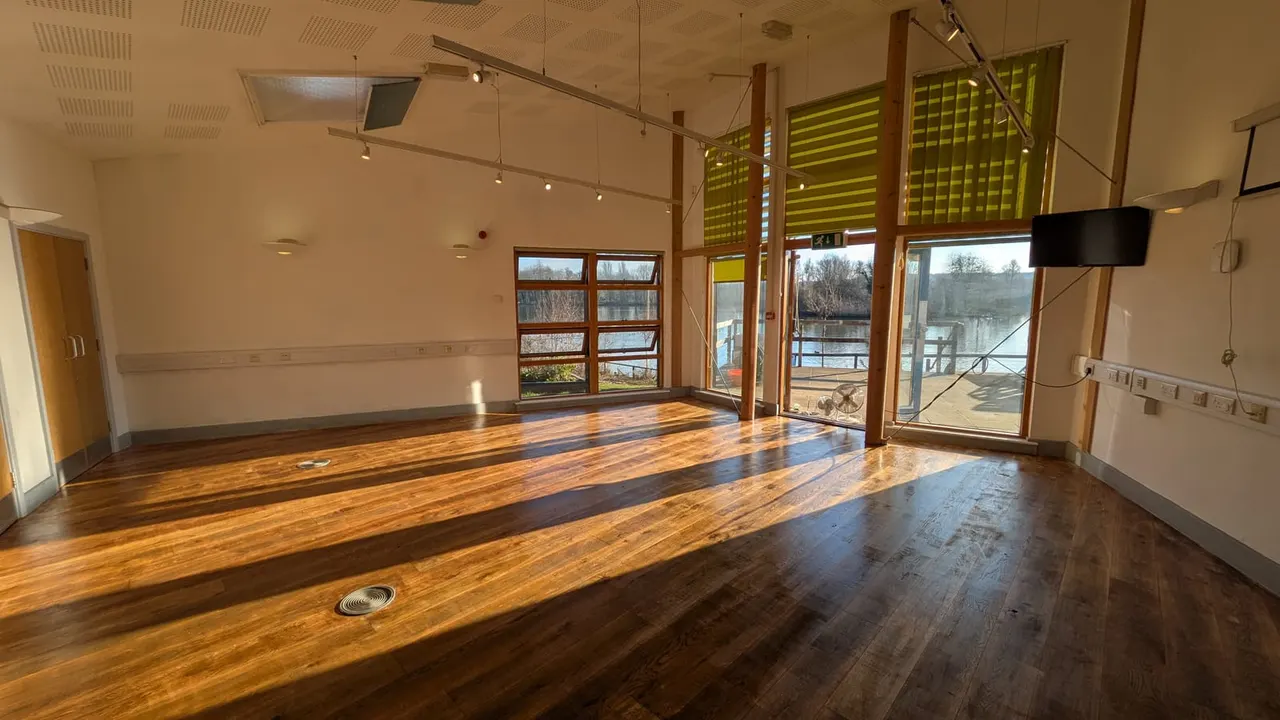 Spacious indoor hall with wooden flooring, large windows overlooking a lake, and green louvred screens, suitable for events and activities.