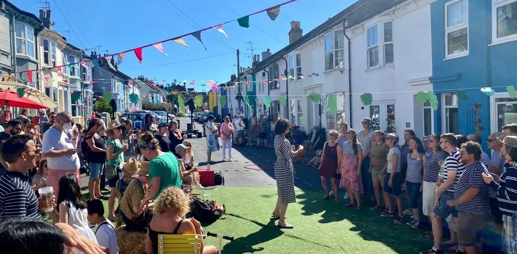 A large crowd of people gathered on a sunny high street lined with colourful buildings, bunting overhead, and a red parasol visible on the left.