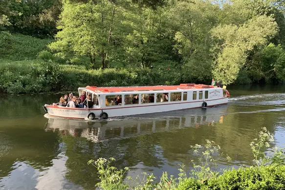A red and white passenger boat carrying tourists cruises along a tree-lined river.