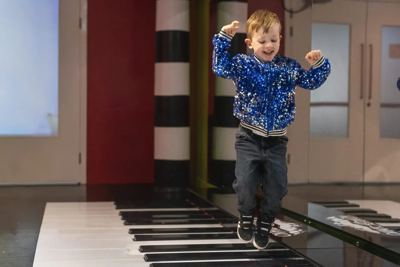 Young child in blue sequined jacket jumping on giant floor piano keys indoors.