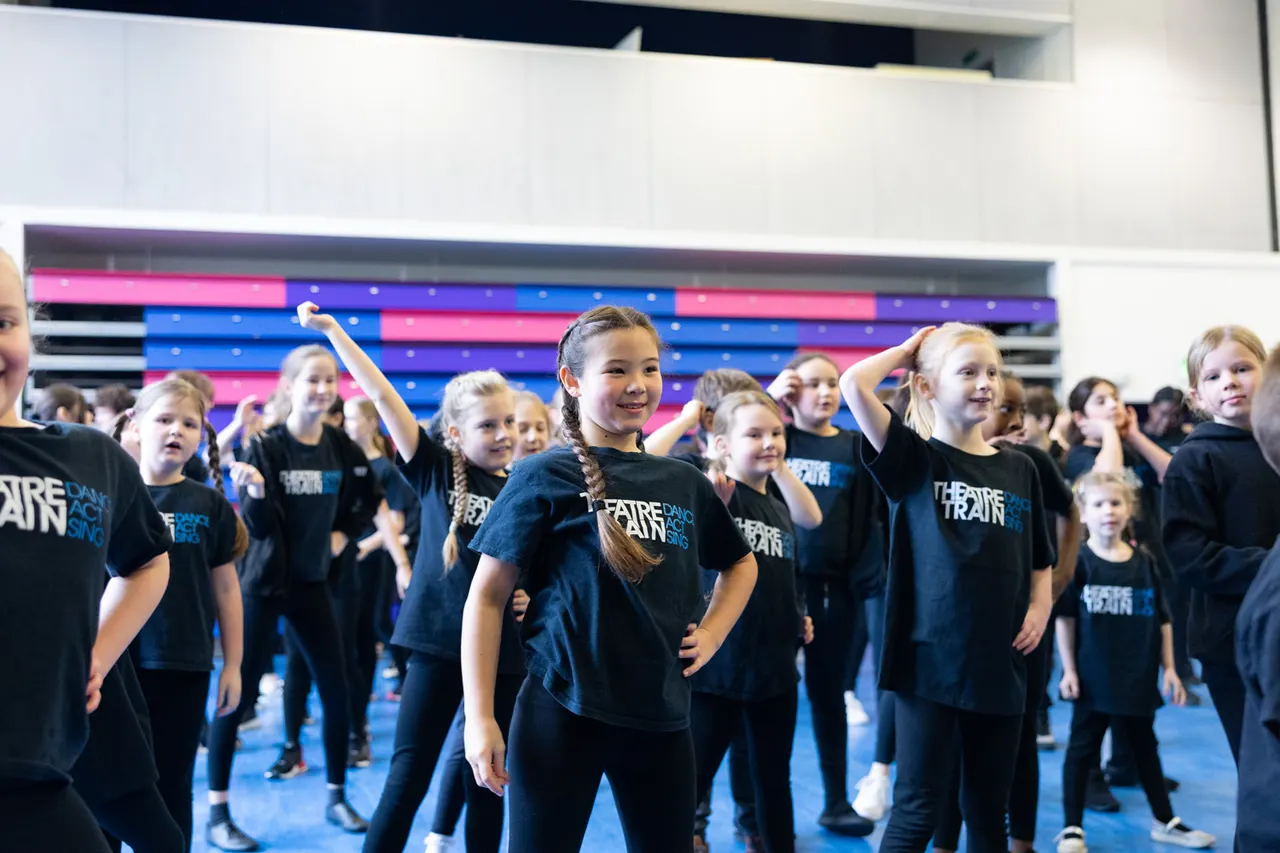 Large group of children in black Theatretrain t-shirts performing on stage with colourful LED backdrop.