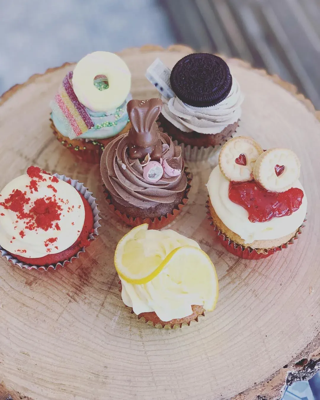 Assorted decorated cupcakes arranged on a wooden board, featuring various toppings including biscuits, chocolate, jam and frosting.