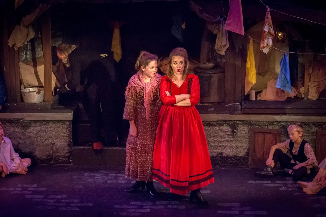 Actors perform a theatrical scene set in a period tenement building with washing lines and makeshift furnishings on stage.