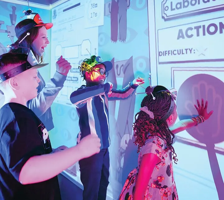 Children wearing VR headsets interact with a glowing interactive display wall in a dimly lit gaming environment.
