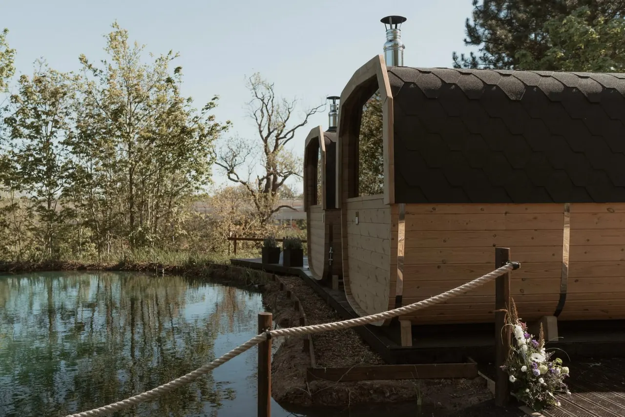 Wooden glamping pods with curved roofs situated beside a still canal, surrounded by trees and connected by a rope barrier.
