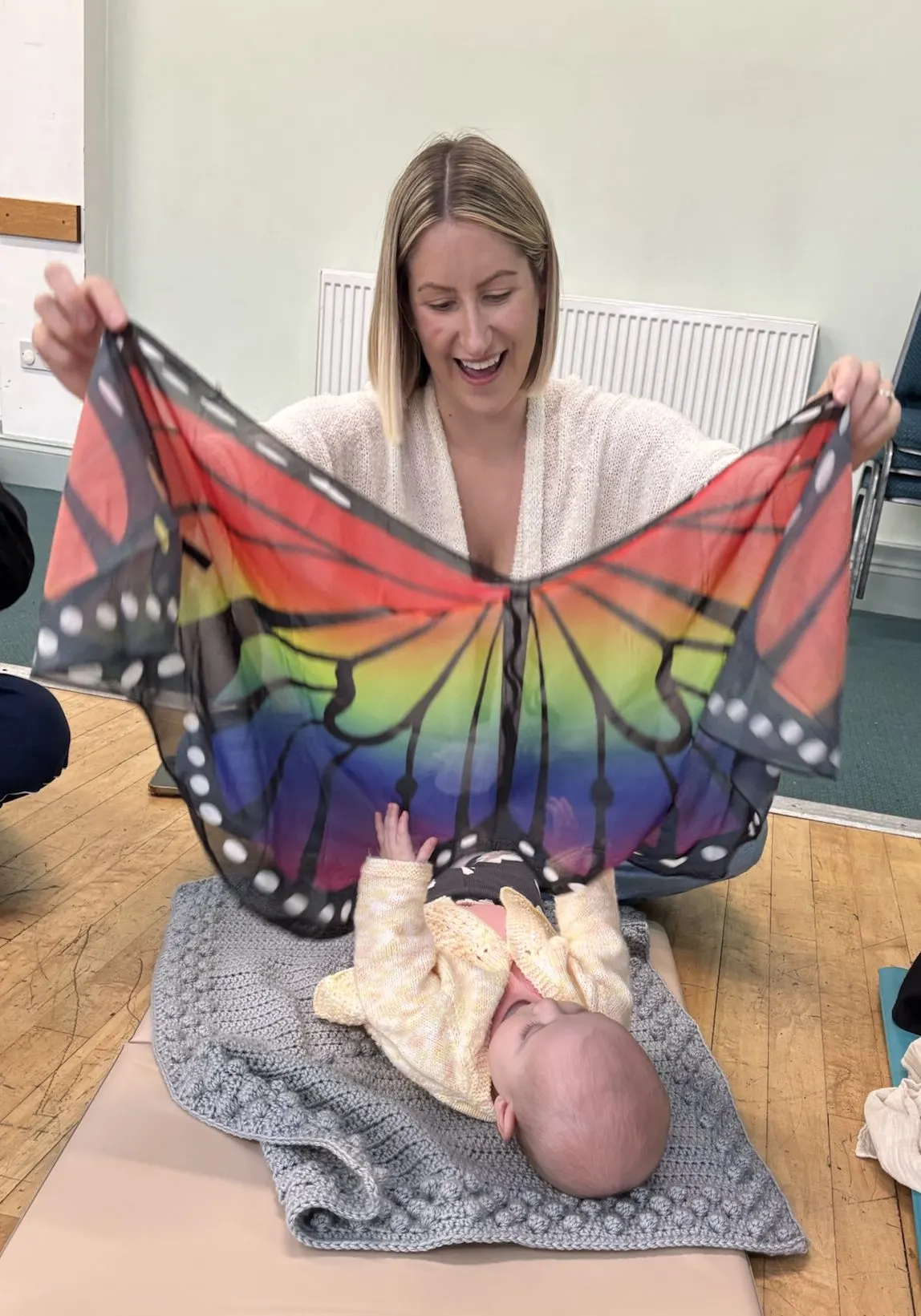 Woman holding a rainbow butterfly fabric above a baby lying on a blanket indoors.