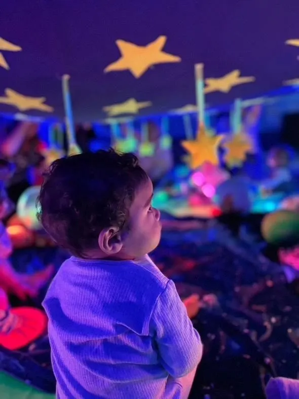 Toddler gazing up at glowing stars and colourful lights in a sensory play environment.