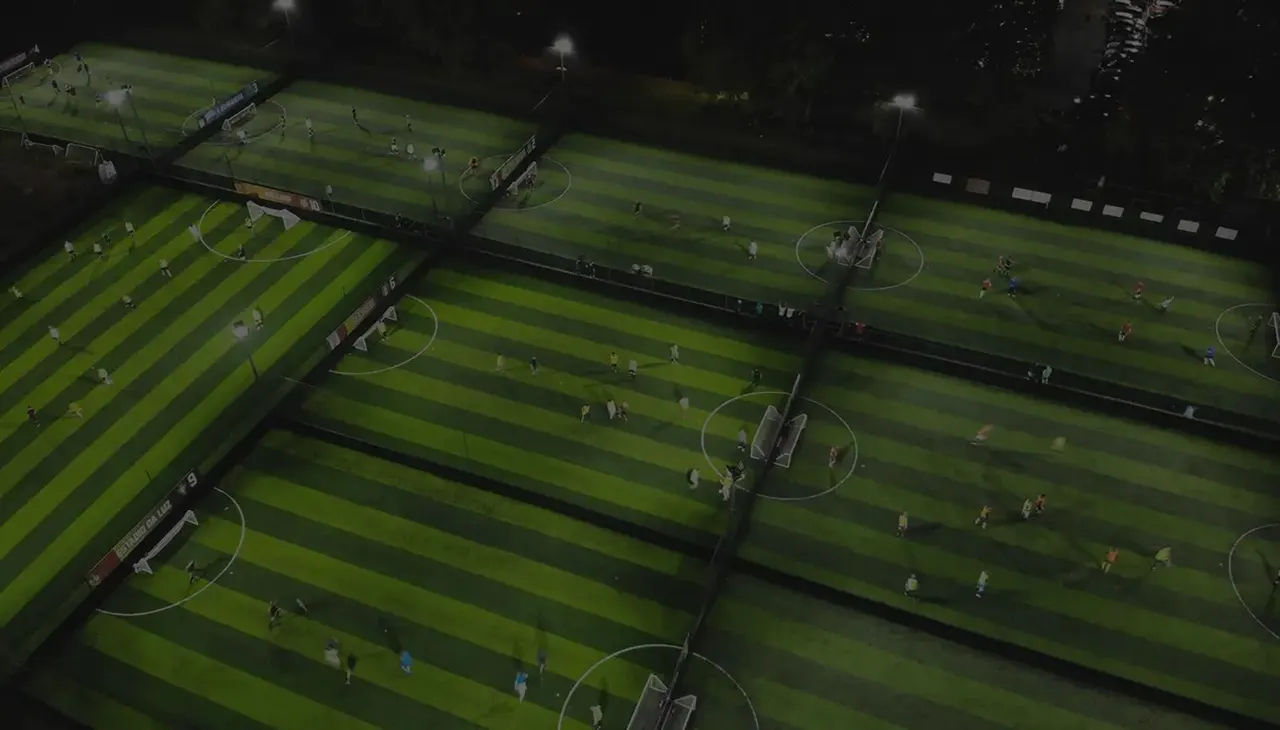 Overhead view of multiple illuminated football pitches with players and small groups scattered across the striped grass during evening play.