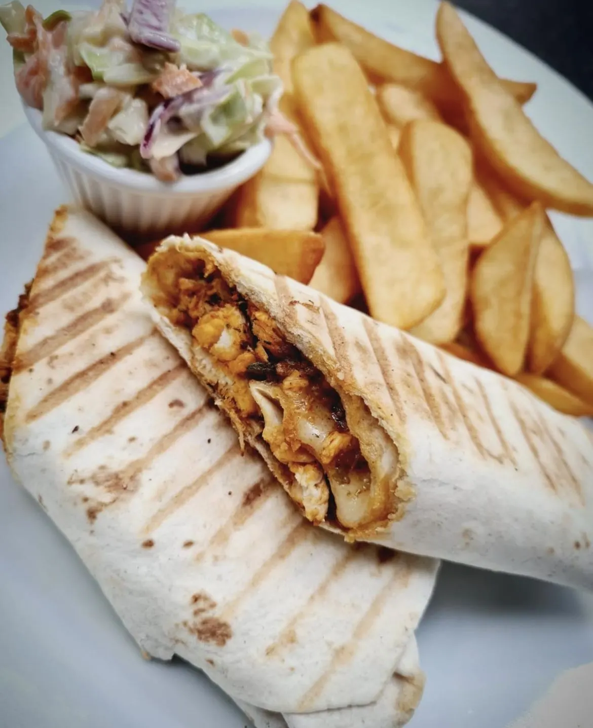 Wrapped sandwich filled with meat and vegetables, served with chips and coleslaw in a small bowl.