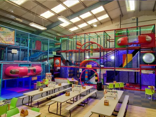 Indoor play area with colourful climbing structures, slides, and tunnels, with picnic tables in the foreground.