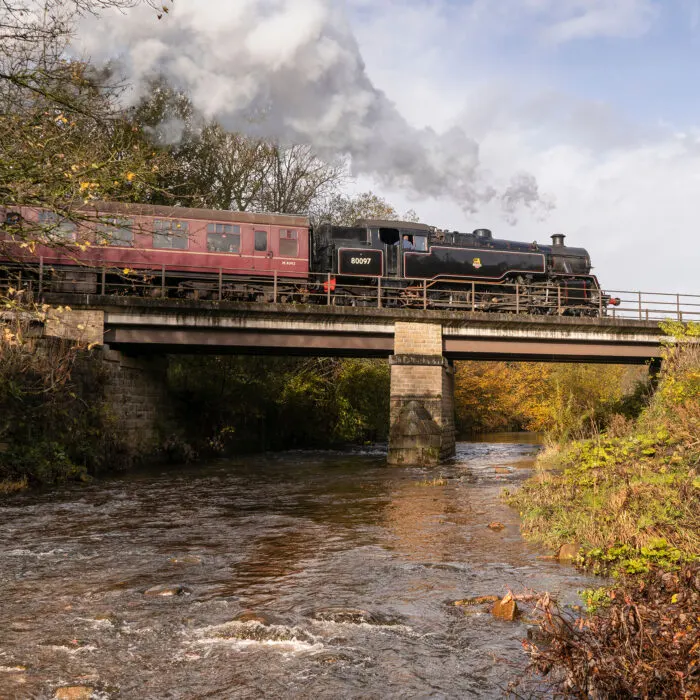Steam locomotives crossing a stone bridge over a flowing river, emitting smoke into the sky.